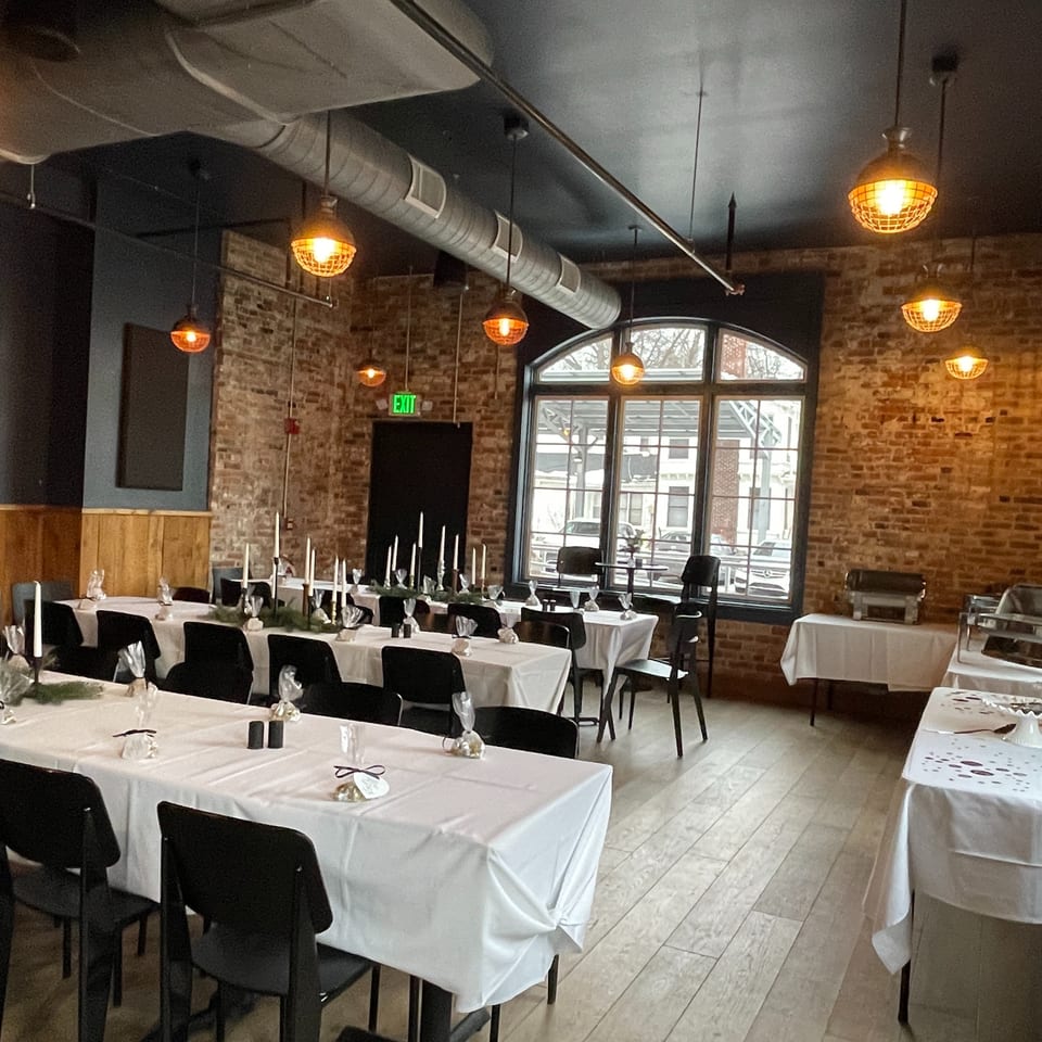 Elegant brick-walled restaurant dining room with white-clothed tables, black chairs, candle centerpieces, and buffet stations.