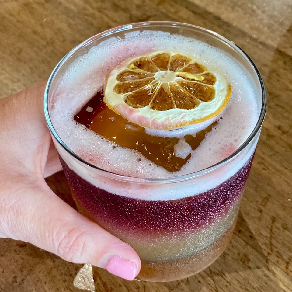 Foamy dark red cocktail garnished with a dried citrus wheel, held in a hand over a wooden table.