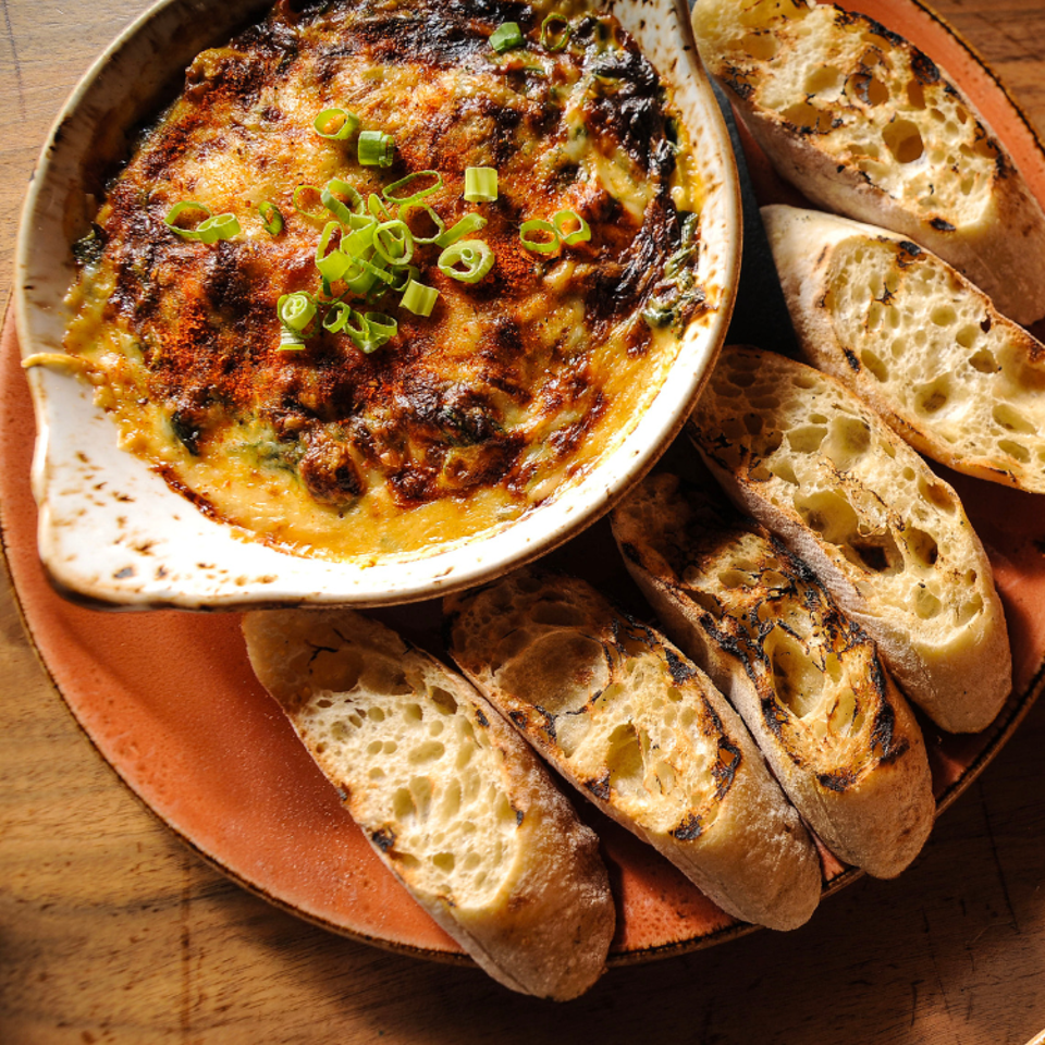 Bubbling baked dip topped with sliced green onions, served in a ceramic dish alongside grilled crusty bread.