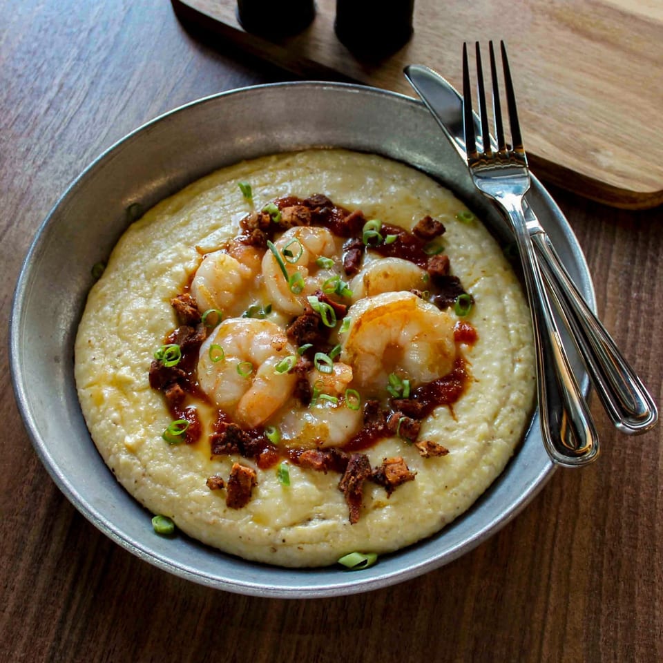 Creamy grits topped with shrimp, crispy bacon, green onions, and sauce in a metal bowl beside fork and spoon.