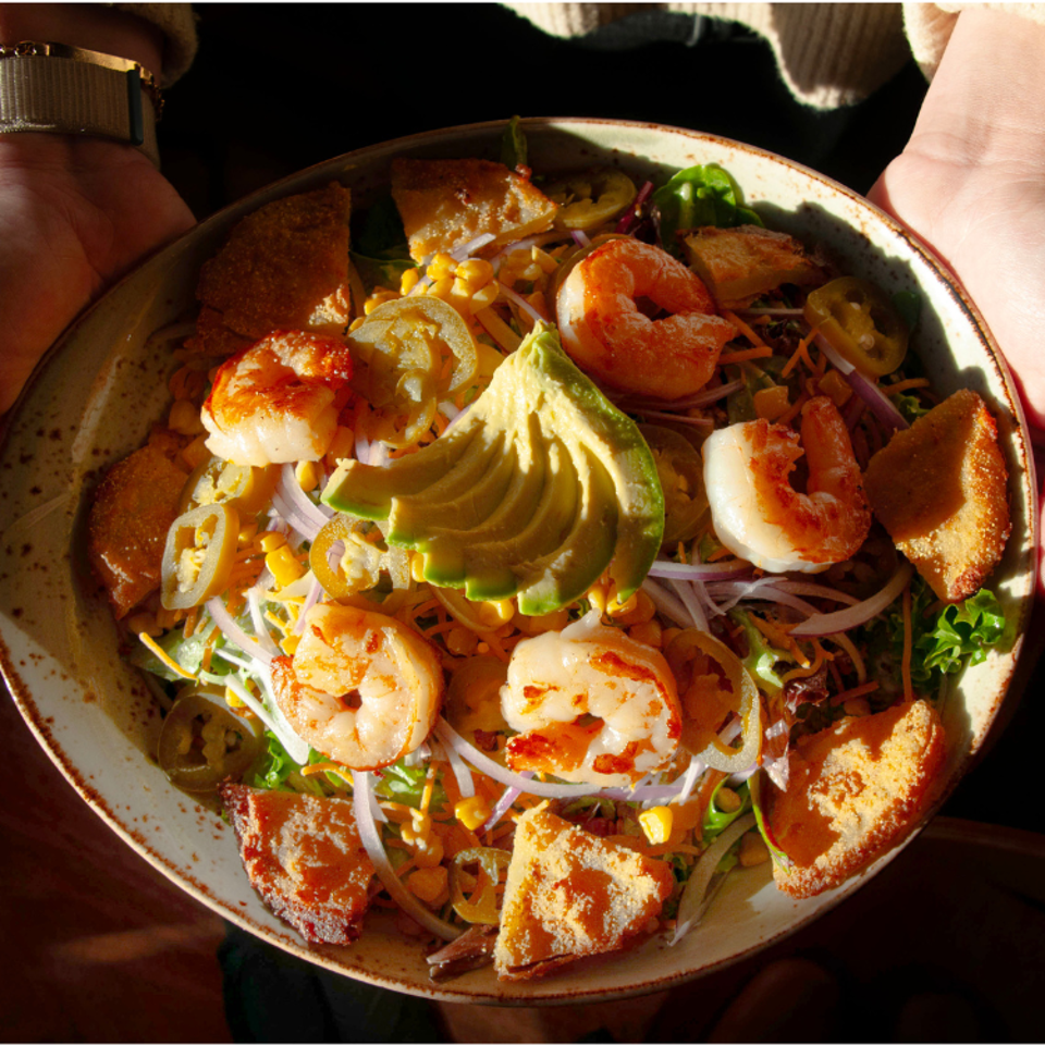 Overhead view of a colorful shrimp salad with avocado, corn, onions, greens, jalapeños, and toasted bread pieces in a bowl.