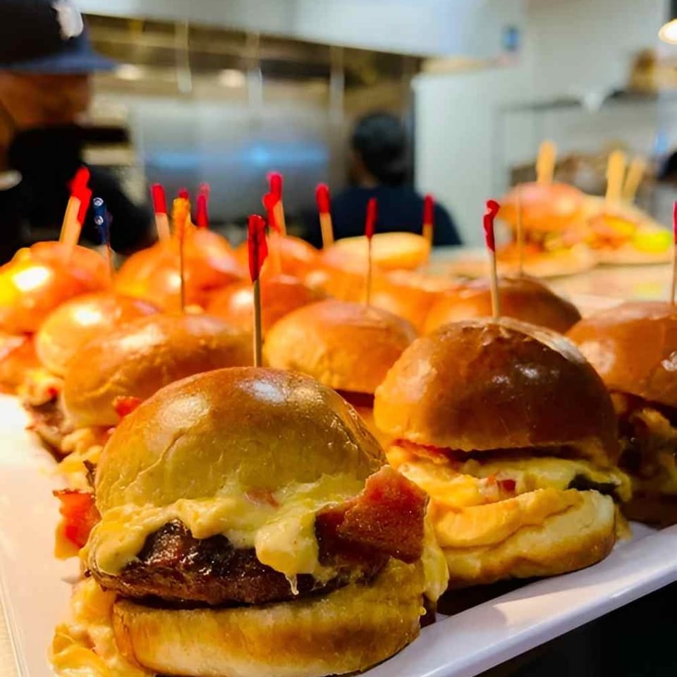 Tray of cheeseburger sliders with glossy buns and toothpicks displayed at a busy restaurant counter.