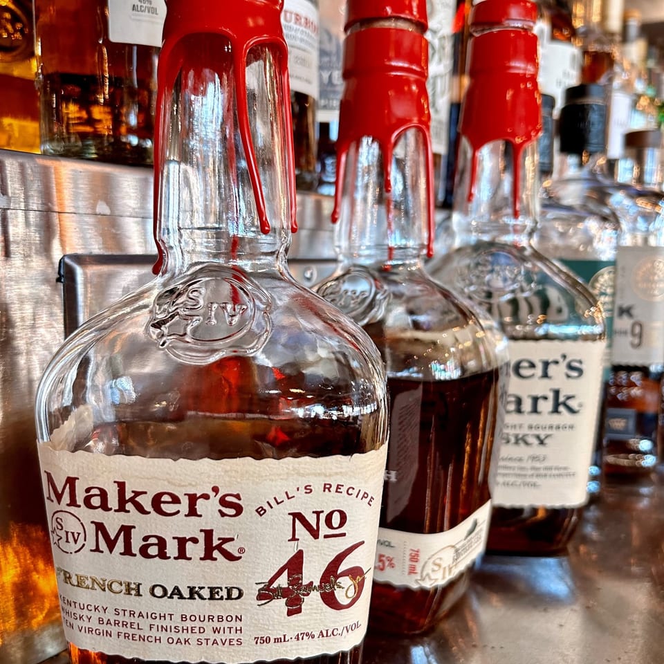 Close-up of Maker’s Mark bourbon bottles with distinctive red wax seals displayed on a warmly lit bar shelf.