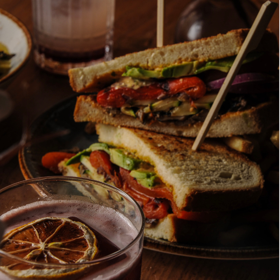 Stacked toasted vegetable sandwiches with avocado, tomatoes, peppers, and onions beside a glass of iced citrus drink.