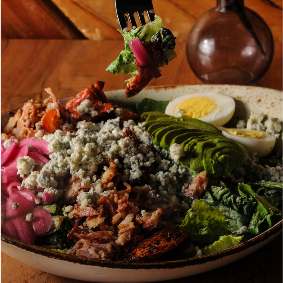 Loaded salad with grilled vegetables, avocado, bacon, blue cheese, pickled onions, and egg on a rustic plate.