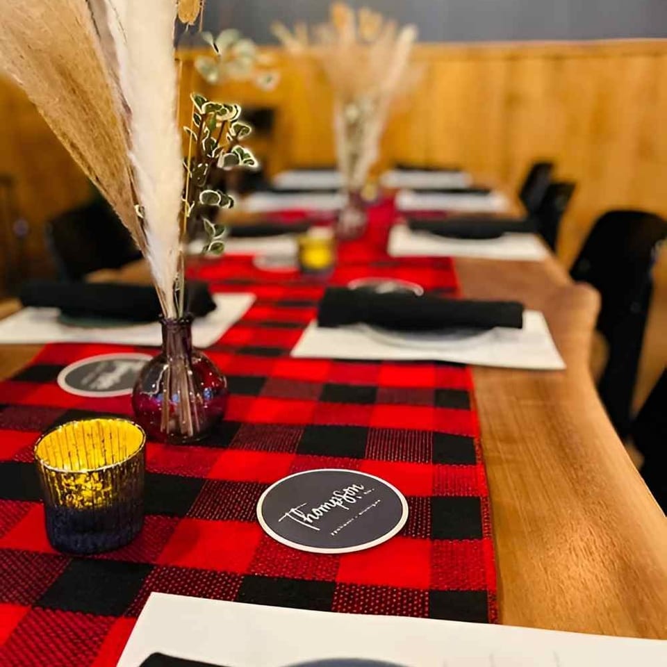 Restaurant table set with red-and-black plaid runner, black napkins, plates, decorative dried flowers, and candlelight.
