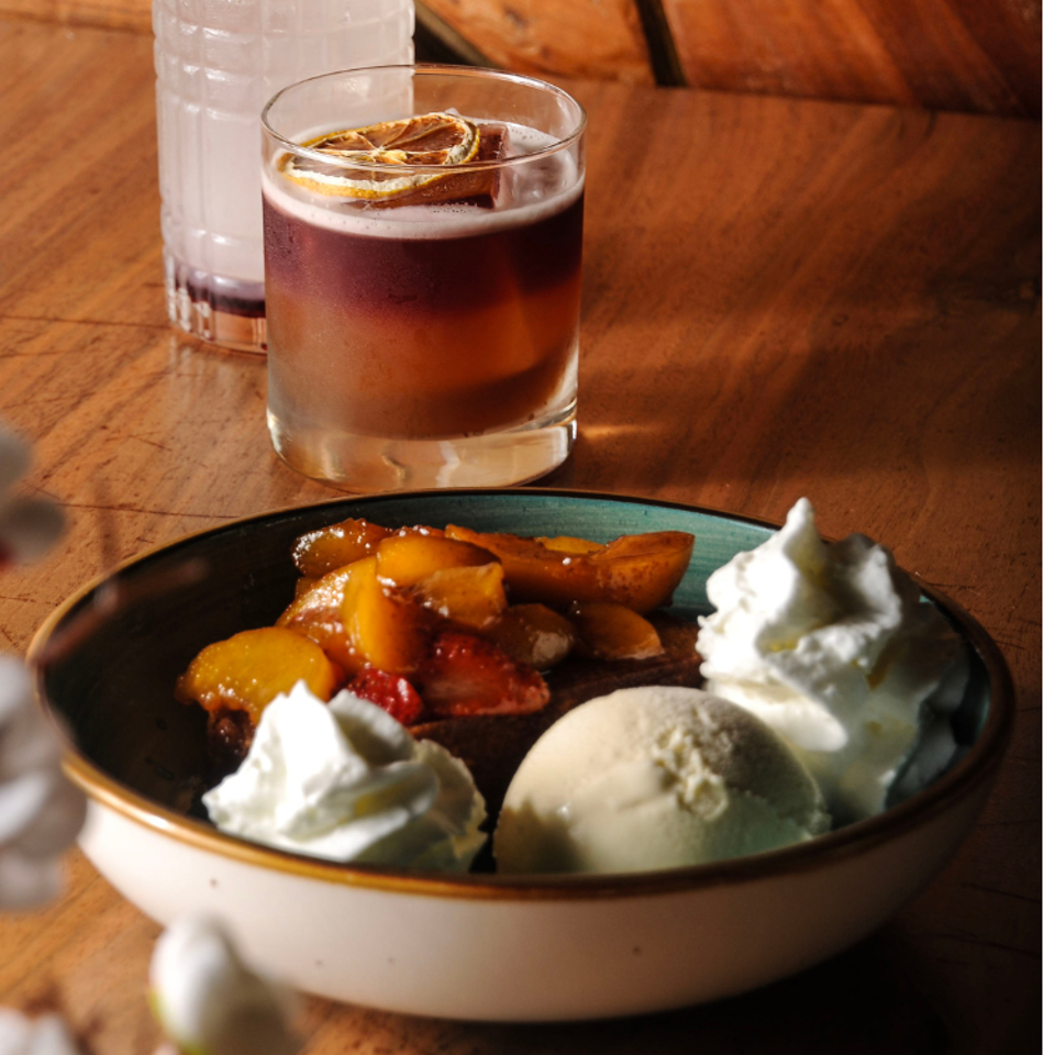 Dessert bowl with roasted fruit, vanilla ice cream, and whipped cream beside a dark citrus-garnished cocktail on a wooden table.