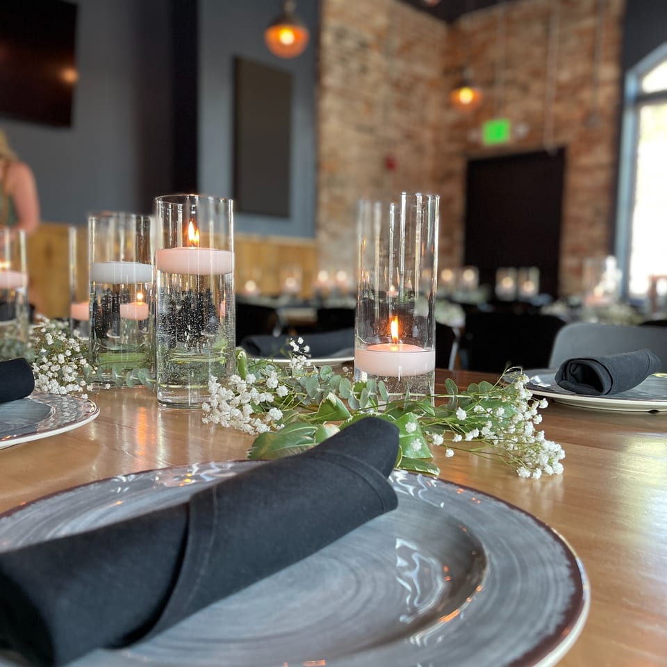 Elegantly set restaurant table with gray plates, black napkins, candlelit glass cylinders, and greenery against exposed brick walls.
