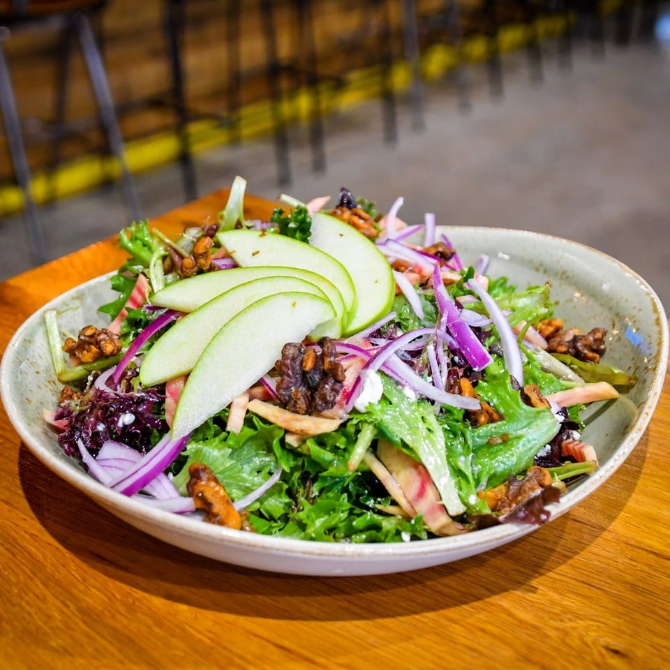 Colorful mixed green salad topped with sliced apples, red onions, radishes, feta, and candied walnuts in a ceramic bowl.