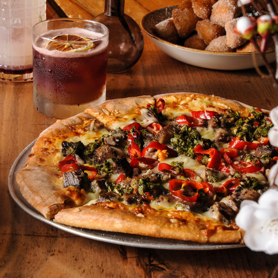 Cheesy vegetable pizza with mushrooms and peppers beside a citrus-garnished drink and powdered pastries on a wooden table.