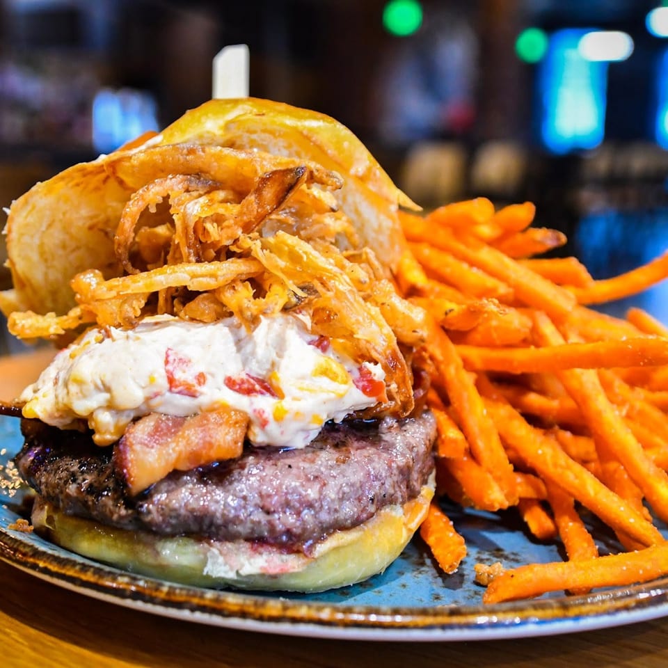 Loaded burger topped with crispy onion rings, bacon, and creamy sauce beside sweet potato fries on a plate.