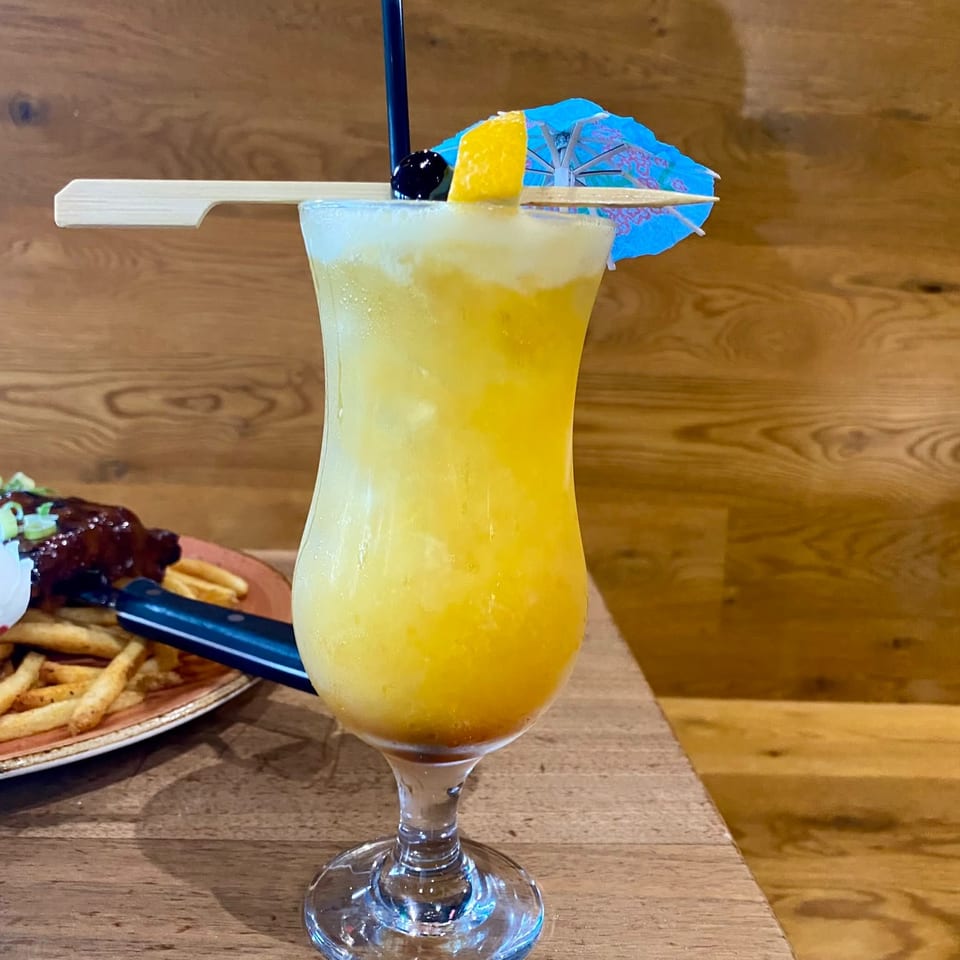 Yellow tropical cocktail garnished with lemon, cherry, straw, and blue paper umbrella beside fries on a wooden restaurant table.