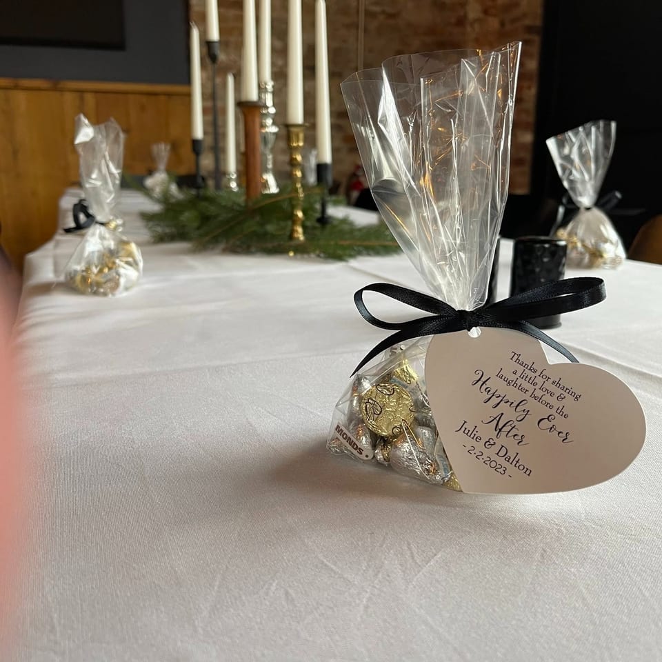 Wedding reception table with wrapped chocolate favors, black ribbons, white linens, greenery, and tall candles against a brick wall.